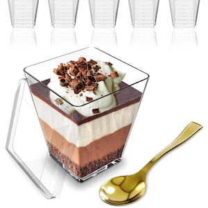 May include: Clear plastic dessert cups and a gold spoon are displayed. One cup is filled with a layered dessert of chocolate, cream, and crumbs, topped with chocolate shavings. The other cups are empty.