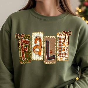 May include: An olive green crewneck sweatshirt featuring the word FALL in large, blocky letters. Each letter has a unique pattern, including plaid, leopard print, and autumn-themed motifs, with small decorative bows on the F and L letters.