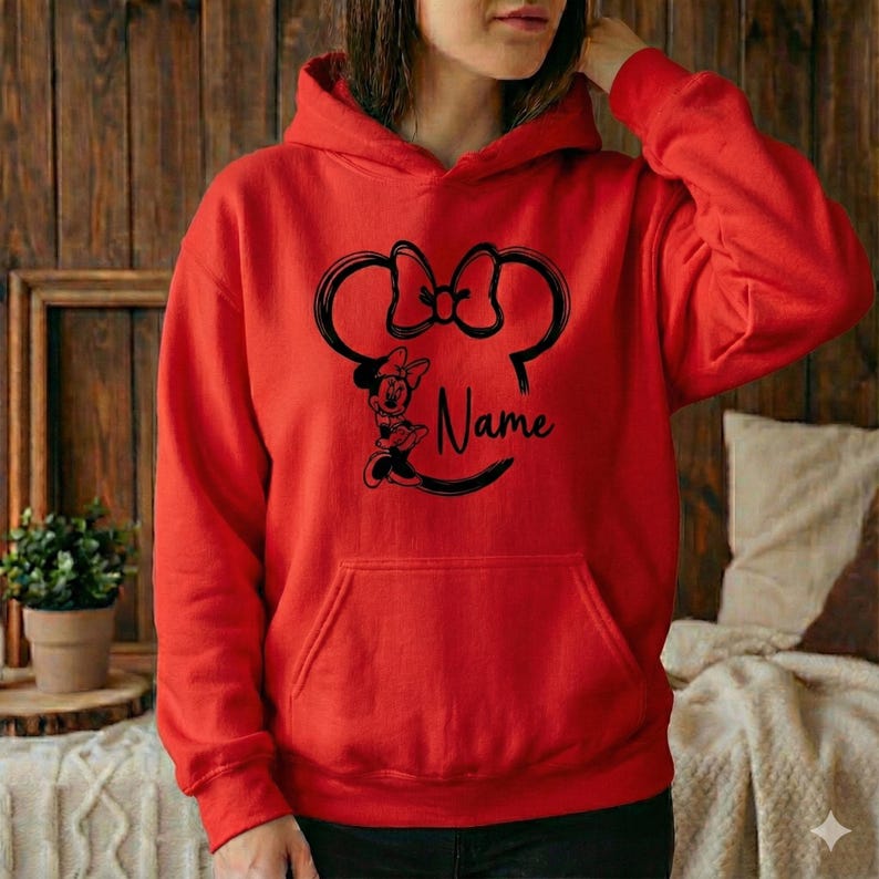 Pu&ograve; includere: Una felpa con cappuccio rossa con un disegno nero della testa di Minnie Mouse, un fiocco e la parola "Name". La felpa ha una tasca frontale e un cappuccio con coulisse. Lo sfondo &egrave; una parete di legno.
