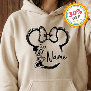 May include: A beige hoodie featuring a black Minnie Mouse design. The design includes Minnie's silhouette, a bow, and the word "Name" in script. The hoodie has a front pocket and a hood.