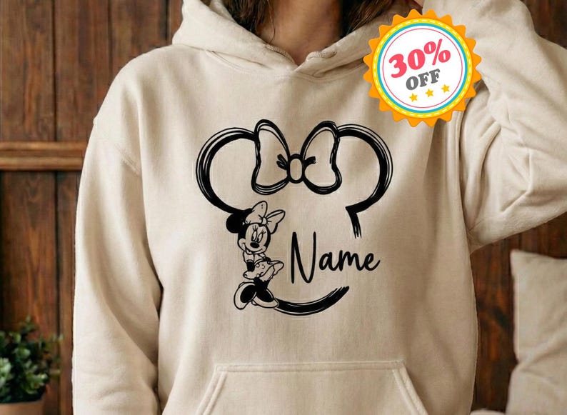 May include: A beige hoodie featuring a black Minnie Mouse design. The design includes Minnie's silhouette, a bow, and the word "Name" in script. The hoodie has a front pocket and a hood.