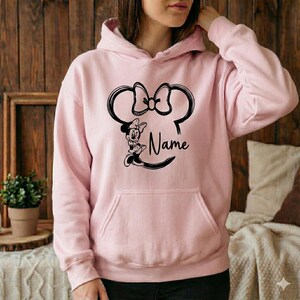 Pu&ograve; includere: Felpa con cappuccio rosa chiaro con un disegno nero di orecchie e fiocco di Minnie Mouse, con la parola "Name" sotto. La felpa ha una tasca a marsupio e un cappuccio con coulisse. Lo sfondo &egrave; un muro di legno.