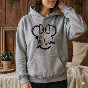 Pu&ograve; includere: Una felpa con cappuccio grigia con una grande grafica nera della testa di Minnie Mouse, il suo fiocco e la parola "Name" in corsivo. La felpa ha una tasca frontale e un cappuccio con coulisse.