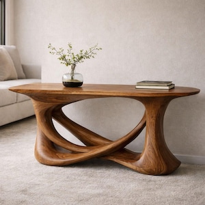May include: A wooden console table with a unique, sculptural base. The table has a natural wood finish and an oval top. A small glass vase with greenery and a stack of books are on the table. The table is approximately 60 inches long.