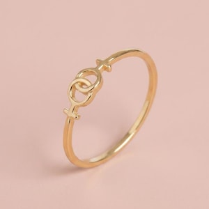 May include: A thin gold ring featuring two interlocking female gender symbols. The band is smooth and circular, with the two connected symbols positioned at the top center of the ring, creating a continuous and delicate metallic design.