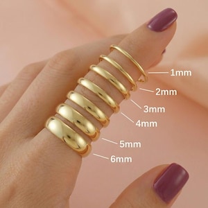 May include: A set of six smooth, polished gold-tone rings stacked on a finger, with labels indicating widths of 1mm, 2mm, 3mm, 4mm, 5mm, and 6mm. Each ring features a simple, rounded band design with a reflective metallic finish and no additional ornamentation.