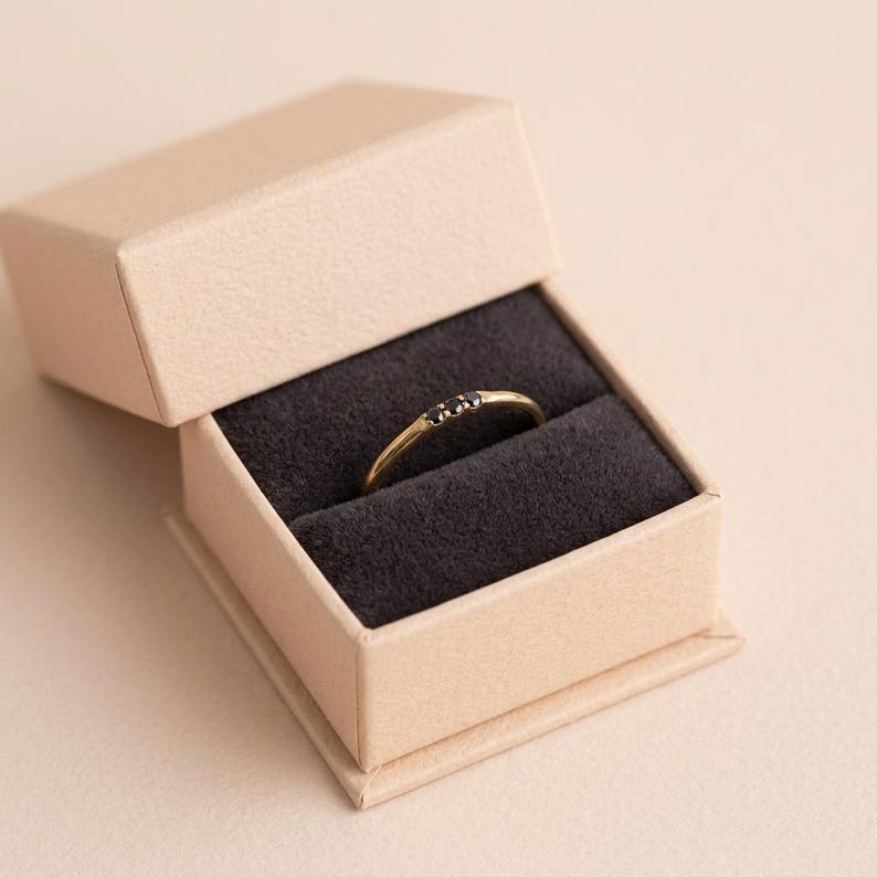 Op de afbeelding: Een dunne gouden ring met drie kleine, ronde zwarte stenen op een rij boven op de band. De ring wordt gepresenteerd in een open, lichtbruin vierkant juwelendoosje met een donkergrijze fluwelen inzetstuk om de ring op zijn plek te houden.