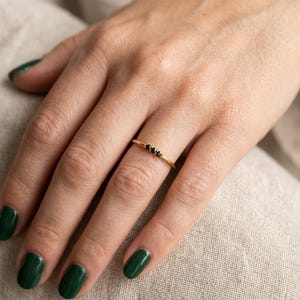 Op de afbeelding: Een dunne gouden ring met drie kleine, ronde, zwarte stenen die in een horizontale rij in het midden zijn geplaatst. De ring heeft een gepolijste afwerking en een eenvoudig, slank ontwerp dat comfortabel om de vinger zit en een strakke uitstraling heeft.