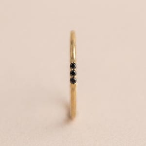 Op de afbeelding: Een dunne gouden ring met een getextureerde band en drie kleine, ronde, zwarte stenen die verticaal op een rij zijn gezet. Het metaal heeft een licht gehamerde of organische afwerking en de stenen worden vastgehouden door fijne metalen pootjes.