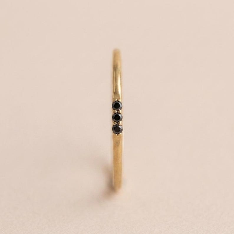Op de afbeelding: Een dunne gouden ring met een getextureerde band en drie kleine, ronde, zwarte stenen die verticaal op een rij zijn gezet. Het metaal heeft een licht gehamerde of organische afwerking en de stenen worden vastgehouden door fijne metalen pootjes.