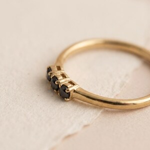 Op de afbeelding: Een gouden ring met drie kleine, ronde, zwarte stenen op een rij. De band is glad en gepolijst met een dun, afgerond profiel. De zetting houdt de drie donkere edelstenen stevig op hun plek, wat zorgt voor een eenvoudig en strak ontwerp.
