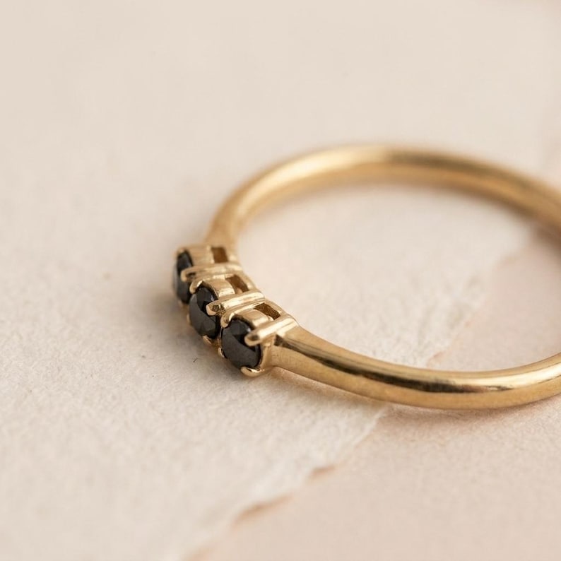Op de afbeelding: Een gouden ring met drie kleine, ronde, zwarte stenen op een rij. De band is glad en gepolijst met een dun, afgerond profiel. De zetting houdt de drie donkere edelstenen stevig op hun plek, wat zorgt voor een eenvoudig en strak ontwerp.
