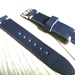 May include: A dark blue leather watch strap with white stitching and a silver buckle. The strap has five holes for adjusting the fit.