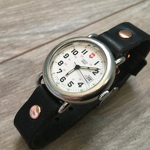 Puede incluir: Un reloj de pulsera de acero inoxidable Swiss Army con esfera blanca y números negros. El reloj tiene una correa de cuero negro con una hebilla de cobre.