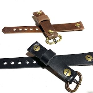 May include: Two leather watch straps, one brown and one black, with brass buckles and screws. The straps have multiple holes for adjusting the fit.
