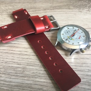 May include: A red leather watch strap with white stitching and a silver buckle. The strap has six holes for adjusting the fit. The strap is shown with a silver chronograph watch with a white face.