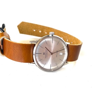 May include: A silver wristwatch with a white face and black hands. The watch has a brown leather strap and a date window at the 3 o'clock position. The watch face has the text "Hamilton Automatic" printed on it.