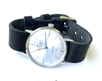 May include: A Hamilton Intra-matic automatic wristwatch with a silver dial, thin black hour markers, and a date window at the six o'clock position. The watch features a polished silver-tone case and a wide, matte black leather strap with a buckle.