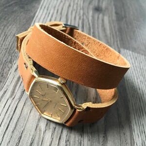 May include: A brown leather watch strap wrapped around a gold watch with a square face.