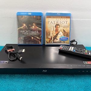 May include: A black LG Blu-ray disc player with a remote control, a black HDMI cable, and two Blu-ray movie cases. The cases feature the titles A Nightmare on Elm Street and The Patriot. A small sticker with the phone number 1-800-243-0000 is visible.