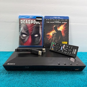 May include: A black Sony Blu-ray disc player with a remote control, a high-speed HDMI cable, and two movie cases: Deadpool and The Dark Knight Rises. The player features a front-loading disc slot, a USB port, and several control buttons.