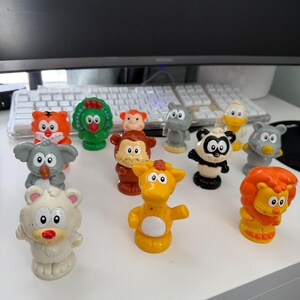 May include: A collection of twelve small plastic animal figurines including a tiger, turkey, monkey, hippo, duck, koala, panda, bear, giraffe, and lion. Each figure features a cartoon-style design with large white eyes and distinct colors.