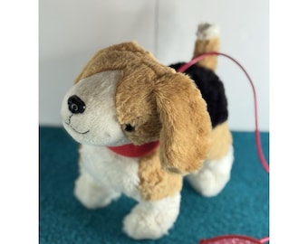 Peut inclure: Un chien en peluche de type beagle avec une fourrure beige, blanche et noire, doté d'un collier rouge et d'un nez en plastique noir. Le jouet est attaché à une laisse rétractable rose avec une poignée affichant le texte KID en blanc.
