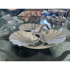 Könnte beinhalten: Eine silberfarbene, muschelförmige Servierschale mit Griff. Die Schale hat zwei Fächer und steht auf einem Perlen-Platzset. Der Griff ist gebogen und scheint aus dem gleichen Metall wie die Schale zu sein.