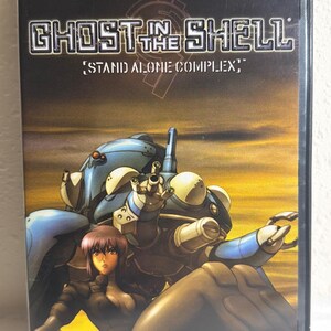May include: A DVD case for Ghost in the Shell: Stand Alone Complex Vol. 02. The cover features a purple-haired character in a dark outfit standing in front of a large blue and white robotic tank against a yellow and orange sunset sky background.