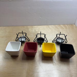Pode incluir: Quatro tigelas quadradas de cerâmica nas cores branca, vermelha, amarela e preta, dispostas em fila. Atrás das tigelas estão três suportes de arame de metal preto, cada um com uma base circular para acomodar uma vela ou aquecedor.