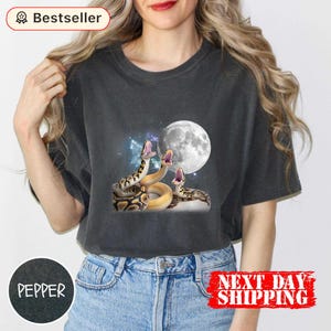 Peut inclure: T-shirt gris foncé avec un motif de trois serpents à gueules ouvertes, une pleine lune et un fond étoilé. Le t-shirt a un col rond et des manches courtes. Le mot "Bestseller" est dans le coin supérieur gauche.