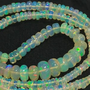 Può includere: Primo piano di più fili di collane di perline di opale. Le perline sono traslucide con lampi di colore iridescente, tra cui blu, verde e arancione. Le perline sono infilate insieme, creando un effetto a strati. Lo sfondo è nero.