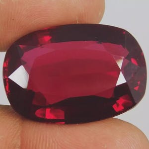 Könnte beinhalten: Ein facettierter, oval geformter Edelstein in tiefroter Farbe. Der Stein hat mehrere Facetten, die das Licht reflektieren und ein brillantes Funkeln erzeugen. Der Stein wird zwischen Fingern gehalten, um seine Größe und Farbe zu zeigen.