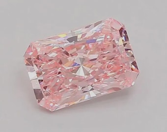 May include: A radiant-cut, light pink diamond. The gemstone has a rectangular shape with multiple facets that reflect light, creating a sparkling effect. The diamond is set against a plain, light background.