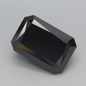 Puede incluir: Una gema negra rectangular facetada, con bordes afilados y superficie pulida. La gema se encuentra sobre un fondo gris claro, resaltando su color oscuro y forma geométrica. El corte crea múltiples superficies reflectantes.