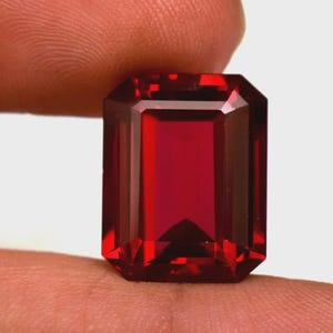 Può includere: Una gemma rosso scuro, tagliata a forma rettangolare e sfaccettata. La gemma ha una superficie brillante e lucida con bordi netti e un colore ricco e saturo. La gemma è tenuta tra due dita.
