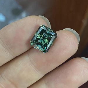 Pode incluir: Uma pedra preciosa retangular, de cor verde-azulado, com lapidação radiante. A gema exibe múltiplas facetas, criando uma exibição brilhante de luz e cor. A pedra é segurada entre os dedos, mostrando seu tamanho e clareza.