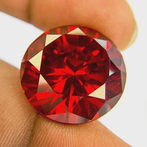 Può includere: Una gemma rosso scuro, tagliata a forma rotonda e sfaccettata. La gemma è tenuta tra le dita, mettendo in risalto il suo taglio brillante e il colore intenso. Le sfaccettature riflettono la luce, creando un effetto scintillante. Adatta per gioielli.