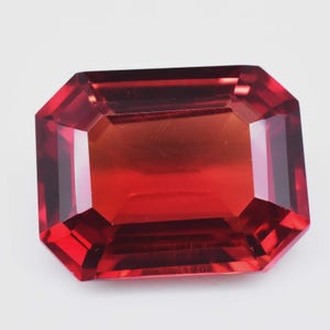 Könnte beinhalten: Ein facettierter, rechteckig geschliffener Edelstein in tiefroter Farbe. Der Stein hat eine polierte Oberfläche und ein symmetrisches Design, das seine Klarheit und Brillanz zeigt. Die Facetten reflektieren das Licht und erzeugen ein lebendiges Farbspiel.