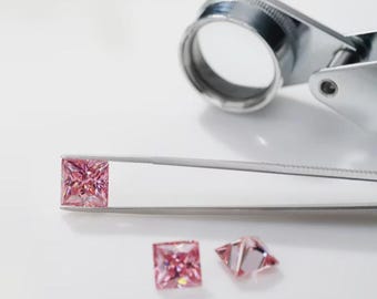 May include: A square-cut, pink gemstone is held by tweezers, with a magnifying glass in the background. Two additional pink gemstones are also visible. The gemstones are set against a white background.