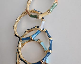 Puede incluir: Brazaletes de metal dorado con detalles de esmalte de colores. Los brazaletes presentan un diseño de bambú con segmentos verdes, blancos, azules y melocotón. Los brazaletes están apilados sobre una superficie blanca.