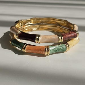 Può includere: Tre braccialetti dorati in stile bambù con segmenti di vari colori. I colori includono sfumature di marrone, verde, arancione e crema, separati da accenti dorati. I braccialetti sono impilati, creando un effetto a strati.