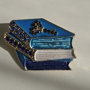 Pode incluir: Broche com uma pilha de três livros em tons de azul e branco, com bordas douradas. Um design de chave adorna o topo. O broche é adornado com strass azuis, adicionando um toque de brilho a este acessório com tema literário.