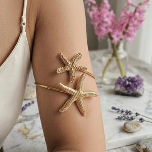 Op de afbeelding: Gouden armmanchet met twee zeesterontwerpen. De armmanchet wordt op de bovenarm gedragen. De zeesterren zijn getextureerd en gedetailleerd, wat een vleugje kustelegantie toevoegt. De sieraden zijn geplaatst tegen een zachte achtergrond met bloemen.