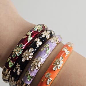 May include: Four colorful enamel bangles with floral designs. The bangles are red, black, lavender, and orange, each adorned with white and gold flowers and green leaves. The bracelets have a gold-colored metal base.
