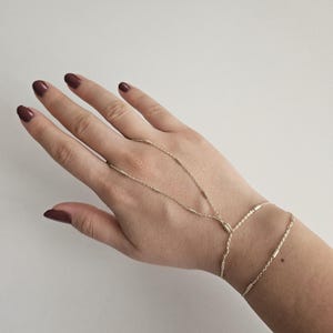 Puede incluir: Una delicada pulsera de cadena de mano dorada. La pulsera presenta una cadena que se conecta a un anillo y una pulsera que se envuelve alrededor de la muñeca. La joya se muestra en una mano con esmalte de uñas rojo oscuro.