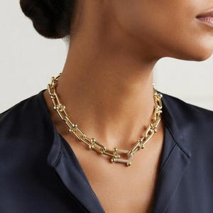 May include: A gold-toned chain necklace featuring elongated oval links with small spherical accents at each joint. The necklace includes a rectangular central link encrusted with small, clear, sparkling stones, serving as a focal point.