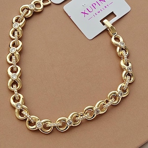 Puede incluir: Pulsera dorada con un diseño de eslabones repetidos, adornada con pequeñas gemas transparentes. La pulsera se muestra sobre un fondo neutro, con una etiqueta blanca que dice "XUPING - JEWELRY-".