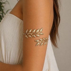 May include: Gold-toned arm cuff featuring a leaf design. The jewelry piece is worn on the upper arm and has a delicate, nature-inspired aesthetic. The cuff is made of metal and has a shiny finish.