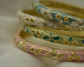 Könnte beinhalten: Vier gestapelte Armreifen mit floralen Mustern. Die goldfarbenen Armreifen sind mit Emaille in Pastellfarben versehen: Rosa, Grün, Weiß und Blau. Jeder Armreif hat ein sich wiederholendes Muster aus Blumen und Blättern mit kleinen Kristallakzenten.